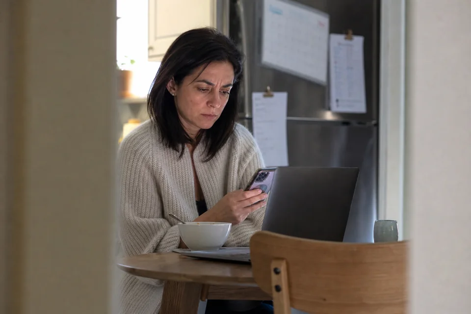 A marketing manager reading a drop alert on her phone beside a laptop at a kitchen table before work